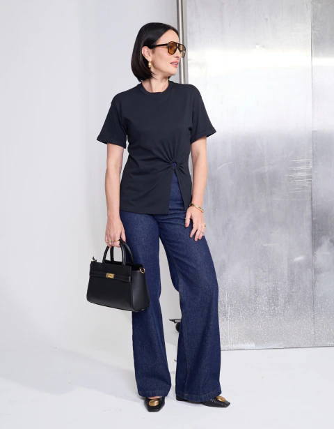 Mulher usando a Camiseta Gaudí Recorte Frontal Preta, combinada com calça jeans de corte reto, segurando uma bolsa preta e usando óculos escuros, em ambiente minimalista.