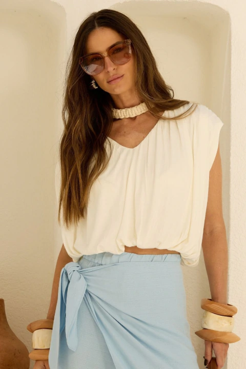 Mulher vestindo blusa de malha franzida off-white, com gola alta e caimento amplo, combinada com saia azul clara e acessórios modernos.
