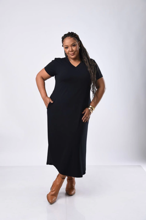 Mulher negra plus size usando vestido malha plus preto de manga curta e comprimento midi, combinando com botas marrons, posando com as mãos nos bolsos.