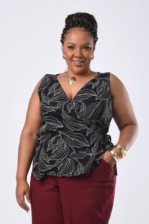 Mulher plus size vestindo blusa transpassada sem manga em tecido preto com estampa de desenho de folhas brancas  com pantalona de linho vinho. 