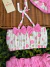 Conjunto infantil de banho de sol com top de bolinhas rosa e bege com laços rosa, calcinha rosa com babados e chapéu rosa, sobre fundo de folhas verdes e superfície de palha