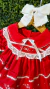 Vestido natalino vermelho para bebê com babados, renda renascença branca e laço de cetim branco no centro do peito, pendurado em cabide de madeira com laço branco na parte superior.