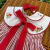 Vestido infantil com listras vermelhas e brancas, gola branca bordada com detalhes de cogumelos e flores, laço vermelho frontal e faixa de cabelo combinando