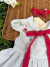 Vestido branco de bebê com bordados delicados de flores e cogumelos, laço vermelho na frente, sobre superfície de vime