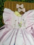 Vestido infantil rosa suave com bordados artesanais de flores e borboletas, mangas amplas com pregas e laços laterais, sobre superfície de vime com dois laços de cabelo combinando