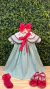 Vestido infantil verde com gola bordada em branco e vermelho, laço vermelho na frente, meia vermelha com laço e sapatos vermelhos brilhantes, fundo de folhas verdes