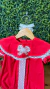Romper natalino vermelho com detalhes em renda branca e laço central, pendurado em cabide de madeira com fundo de plantas verdes