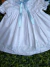 Vestido branco bebê com bordados azuis e laço azul no peito, detalhes em renda nas mangas e gola, tecido leve e delicado