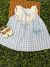 Vestido infantil Helena Xadrez Azul com estampa xadrez em azul claro e branco, gola branca bordada à mão com flores rosas e folhas verdes, laço azul no pescoço e laço azul decorativo na saia, acompanhado por sapatinhos infantis brancos.