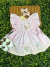 Vestido infantil rosa suave com bordados artesanais de flores e borboletas, mangas amplas com pregas delicadas, laços laterais e saia rodada com acabamento em renda, acompanhado de sapatinhos brancos com laço e dois laços de cabelo rosa claro