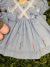 Vestido infantil azul claro com bordados florais delicados e aplicação de renda branca em formato cruzado no corpinho, com mangas bufantes e laços laterais