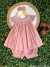 Conjunto infantil rosa estilo batinha com blusinha de ponto smock bordada em rococó, shortinho com elástico e faixa de cabelo combinando, exibido sobre fundo de folhas verdes