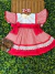 Vestido infantil vermelho com estampa xadrez, detalhes em renda branca, laços laterais e faixa vermelha na cintura, acompanhado de laço de cabelo xadrez e sandálias marrons, sobre fundo de folhagem verde