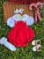 Conjunto infantil com jardineira vermelha, blusa branca com bordado no colarinho, faixa de cabelo branca, meia branca rendada e sapatinhos brancos, sobre folhas verdes e fundo de cesta de vime