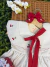 Vestido infantil branco com bordados de cerejas vermelhas e laço vermelho amarrado na frente, exibido em cabide de madeira sobre superfície de vime, com flores brancas ao lado