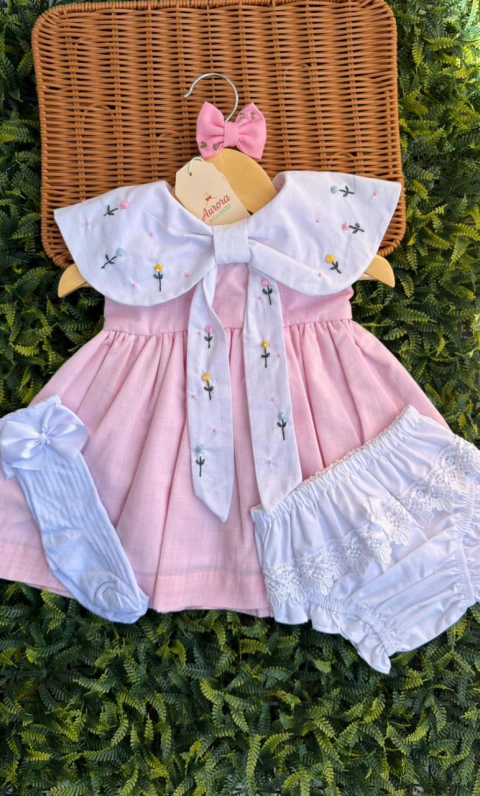 Vestido rosa claro com gola branca bordada com flores coloridas, acompanhado de meia branca e calcinha com renda, sobre fundo de folhagem verde