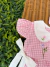 Detalhe de romper infantil rosa xadrez com gola branca e bordados florais, amarração lateral branca, sobre superfície de vime com folhas verdes ao fundo