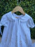 Vestido branco infantil com gola arredondada e renda delicada, com bordados manuais em tom suave e mangas bufantes com laços, pendurado em cabide de madeira