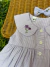 Detalhe da blusa infantil lilás xadrez com gola arredondada e bordado manual de flores roxas, com botões brancos e laço combinando ao lado.