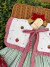 Detalhe do vestido infantil Jardim de Morangos com bordados de morangos vermelhos, babados xadrez vermelhos e listras verdes e brancas.