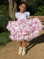 Menina sorridente vestindo vestido Encanto Junino Aurora com estampa colorida de elementos juninos e detalhes em renda, posando ao ar livre segurando a saia ampla