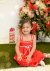 Menina sorridente sentada no chão com vestido vermelho natalino de alcinhas duplas, laço branco e bordados delicados, ao lado de árvore de Natal decorada
