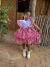 Menina vestindo vestido infantil xadrez vermelho e branco com bordados florais, mangas bufantes e laço lateral, posando em frente a uma casa rústica de barro.