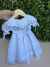 Vestido branco delicado para bebê com detalhes em renda renascença, mangas bufantes e laços laterais, pendurado em cabide de madeira com fundo de folhagem verde