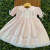 Vestido infantil rosa claro com bordados florais delicados, renda branca e mangas bufantes, pendurado em cabide de madeira sobre fundo de folhagens verdes.