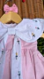 Vestido rosa bebê com gola branca ampla decorada com bordados florais coloridos e laço frontal, pendurado em cabide de madeira com laço rosa