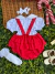 Conjunto infantil de jardineira vermelha com blusa branca de manga curta, meias brancas com detalhes de renda e sapatinhos creme sobre fundo de folhas verdes.