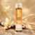 Frasco de Body Splash Warm Vanilla Sugar da Bath & Body Works sobre superfície com fundo bege, decorado com flor de baunilha e favas de baunilha
