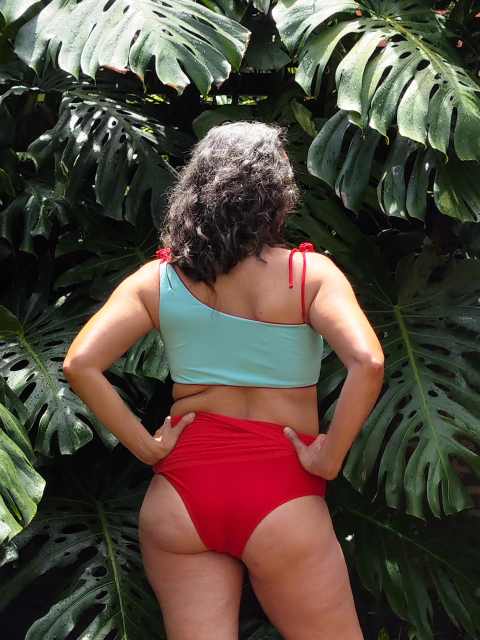 Mujer de espaldas usando un bikini de dos piezas, con parte superior acua y parte inferior roja, rodeada de hojas verdes tropicales.