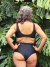 Mujer de espaldas vistiendo un corpiño negro de baño, con plantas tropicales de fondo.