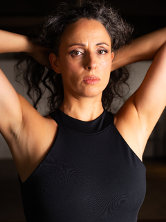 Mujer con cabello rizado oscuro, usando un body negro, con las manos en la cabeza.