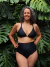 Modelo luciendo un corpiño  triangulo de bikini negro con estampado azul metalizado, combinado con una parte inferior de corte alto, en un entorno natural con plantas verdes.