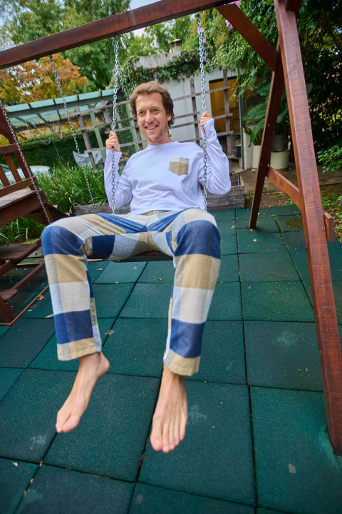 Hombre sentado en un columpio, vistiendo un conjunto de pijama en colores azul y beige, con una sonrisa en su rostro.