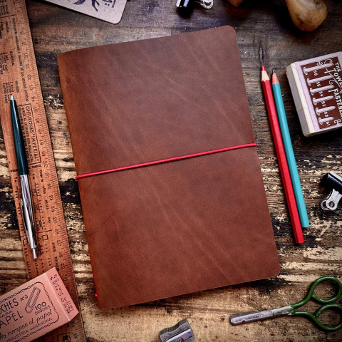 Cubierta de cuero marrón café cerrado con un elástico rojo, sobre una mesa de madera con útiles de papelería