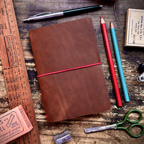 Cubierta de cuaderno PocketCover de cuero color café cerrada, rodeada de útiles de escritorio antiguos sobre una mesa de madera