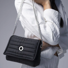 Bolso cruzado negro Andrea Sundar con cadena plateada, presentado en un fondo gris.