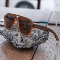 anteojos de sol de madera (patillas) y acetato color marrón rayado oscuro y claro (frente) estilo aviador modelo patagonia marca nomade sobre fondo de madera y piedra