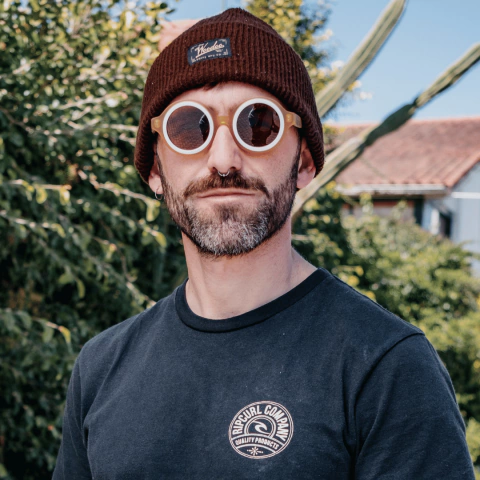 Hombre con anteojos de sol de madera y acetato, usando un gorro marrón, en un entorno natural.