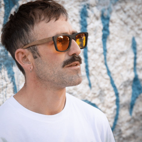 Hombre usando anteojos de madera y acetato Berna TINT, con lentes de color naranja, en un fondo texturizado azul y blanco.