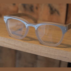anteojos de madera (patillas) y acetato (frente) color cristal con aro de acero inoxidable de forma redondeada para lentes de aumento modelo Dublin marca Nomade sobre repisa de madera