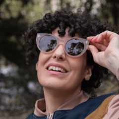 mujer sonriente con anteojos de madera (patillas) y acetato (frente) color rosa traslucido de forma redondeada con lentes polarizados modelo Vera marca Nómade (vista de frente)