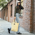 Mujer caminando con una cartera tote de ecocuero color beige, usando un suéter verde y jeans claros, en una calle de ladrillos.