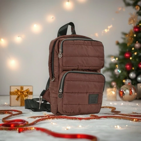 Mochila Hera acolchonada color marrón con dos bolsillos frontales y detalles en cierre plateado, en un entorno navideño con luces y regalos
