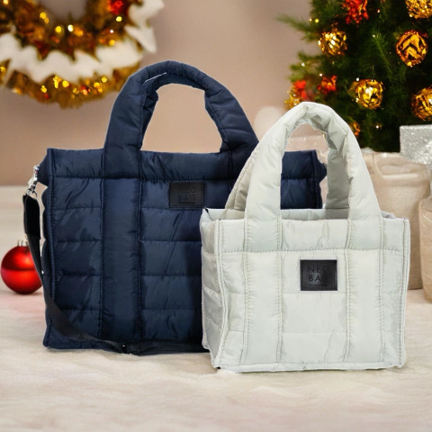Dos tote bags acolchonados de estilo moderno, uno blanco y otro azul oscuro, con asas cortas y etiqueta frontal de cuero, ambientados con decoración navideña de fondo.