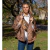Mujer joven con capa acolchonada color chocolate y camisa blanca, posando al aire libre en un parque con árboles