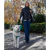 Mujer caminando por un parque en otoño con una capa negra acolchonada y jeans, paseando un perro blanco con arnés verde